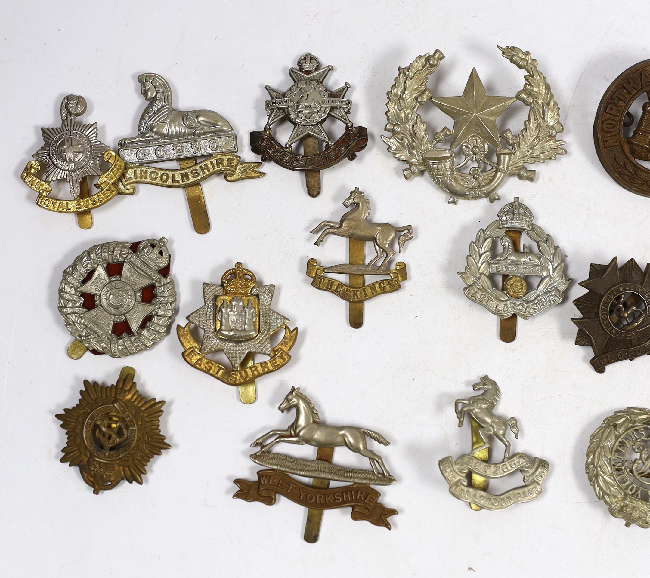 Twenty military cap badges including Bedfordshire, West Yorkshire, Cornwall, East Lancashire, the Royal Scots, the Royal Sussex, Northamptonshire, Lincolnshire, East Surrey, Dorsetshire, Engineer Volunteers, etc.